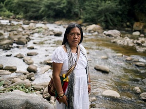 A woman wearing a long white dress with a colourful woven bag on her shoulder stands on the bank of a shallow rocky river  