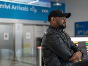 A man wearing a black jacket and cap smiles into the distance in an airport with the Arrivi/Arrivals sign behind him.