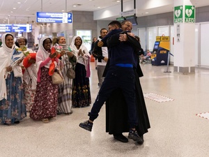 A group of family members embrace tightly at the Athens International Airport, while others stand nearby, smiling and clapping