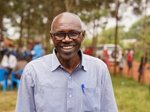 A smiling man wearing a shirt and spectacles stands on a lawn with people and chairs in the background bordered by tall trees