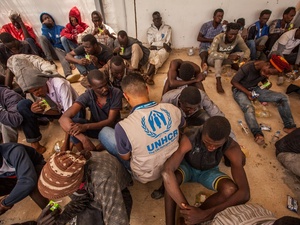 A group of disembarked refugees and migrants sit on the floor, joined by a UNHCR staff member wearing a tan UNHCR vest with blue logo.