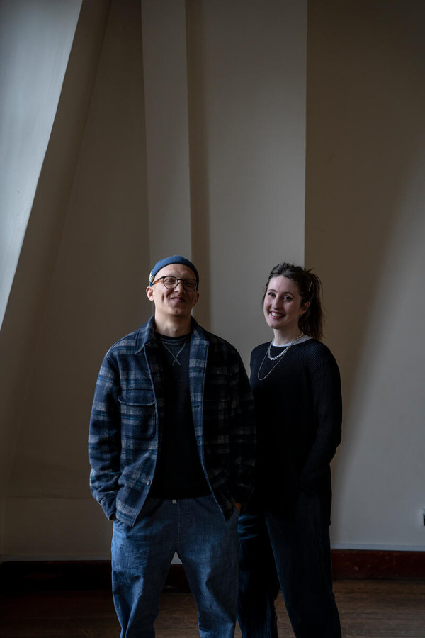 A man and a woman pose standing up against a dark background. 