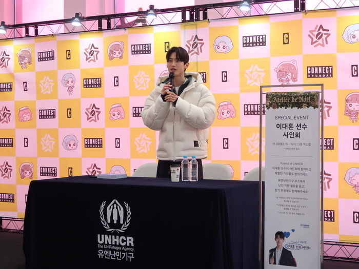Daehoon Lee speaks into a microphone behind a UNHCR Korea-branded table. Behind him is a large banner with Connect Hyundai branding and next to him is a standing banner announcing the special event.