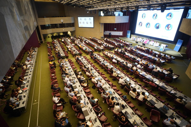 Delegates at rows of desks in a large conference hall listen to a panel discussion on stage, with speaker photos on a big screen.