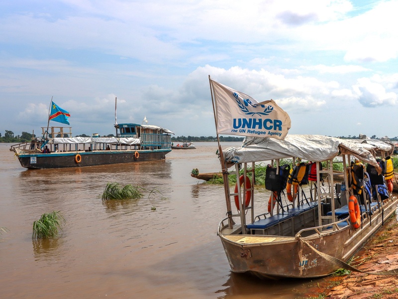 Wooden boats float on a wide, muddy river. One bears a UNHCR flag.