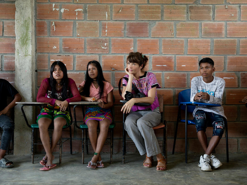 Helena Christensen sits holding a camera with several children at desks against a brick wall