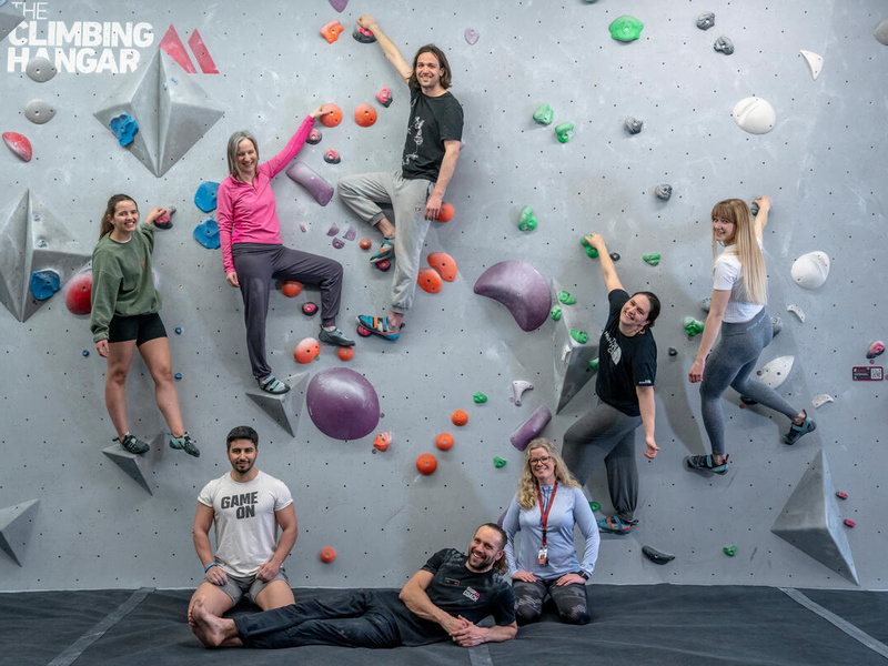 Seven people pose on the floor and on holds on the wall at a bouldering centre.