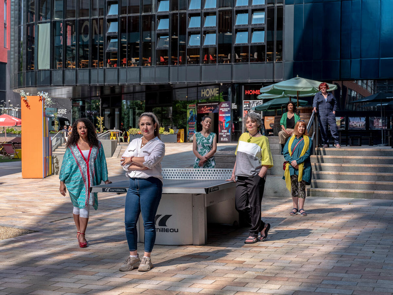Seven women pose for a photo around an outdoor ping pong table.