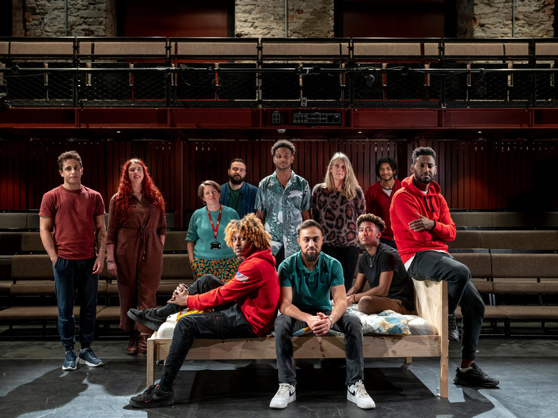 People pose in a theatre, with some sitting on a bed and some standing either side.