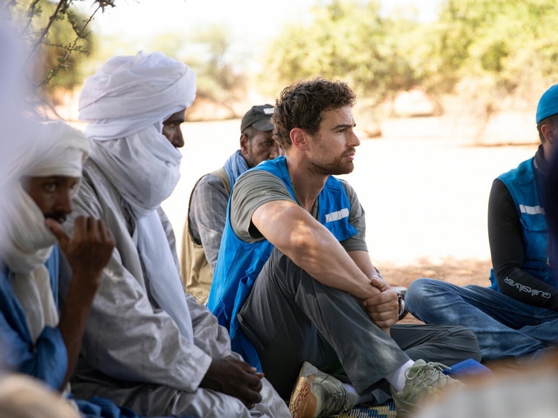 A man wearing a blue UNHCR vest sits on the ground outside next to men wearing white headscarves.