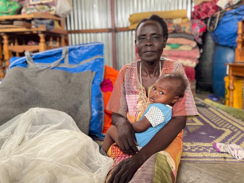 A woman sits on a carpet in an aid distribution center, with a baby on her lap.