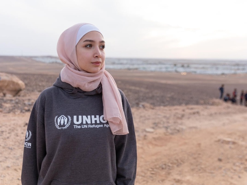 A young woman stands in a desertic area, where a refugee camp is visible in the background