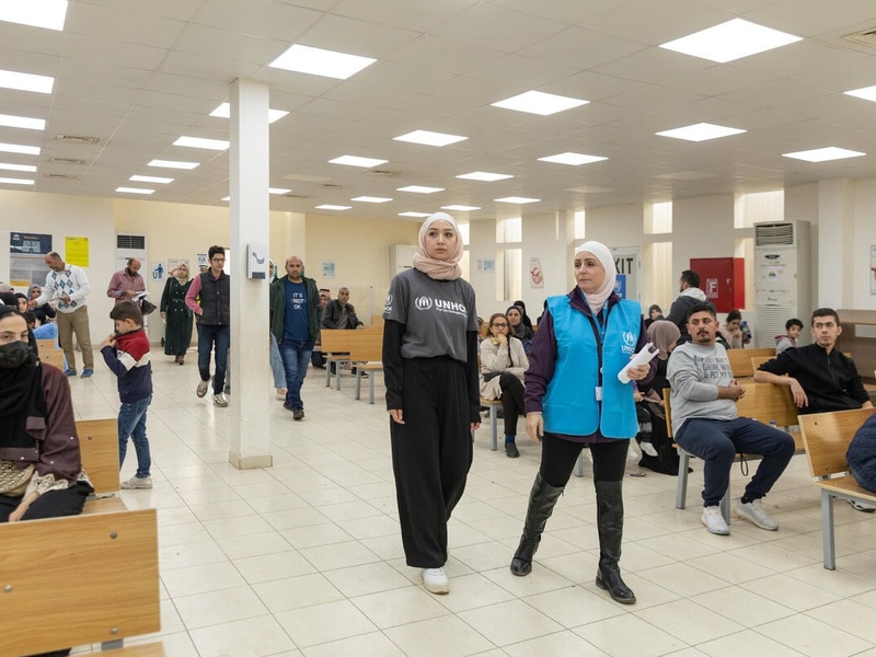Two women walk in a room of the Amman registration center, where other people sit on benches waiting for their turn.