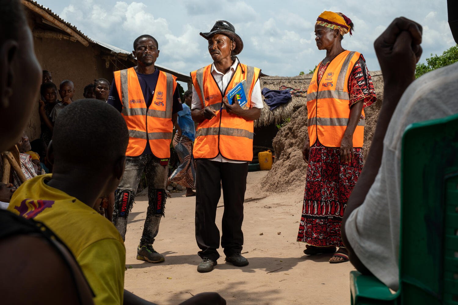 Two men and a woman wearing orange vests talk to an audience sitting outside.