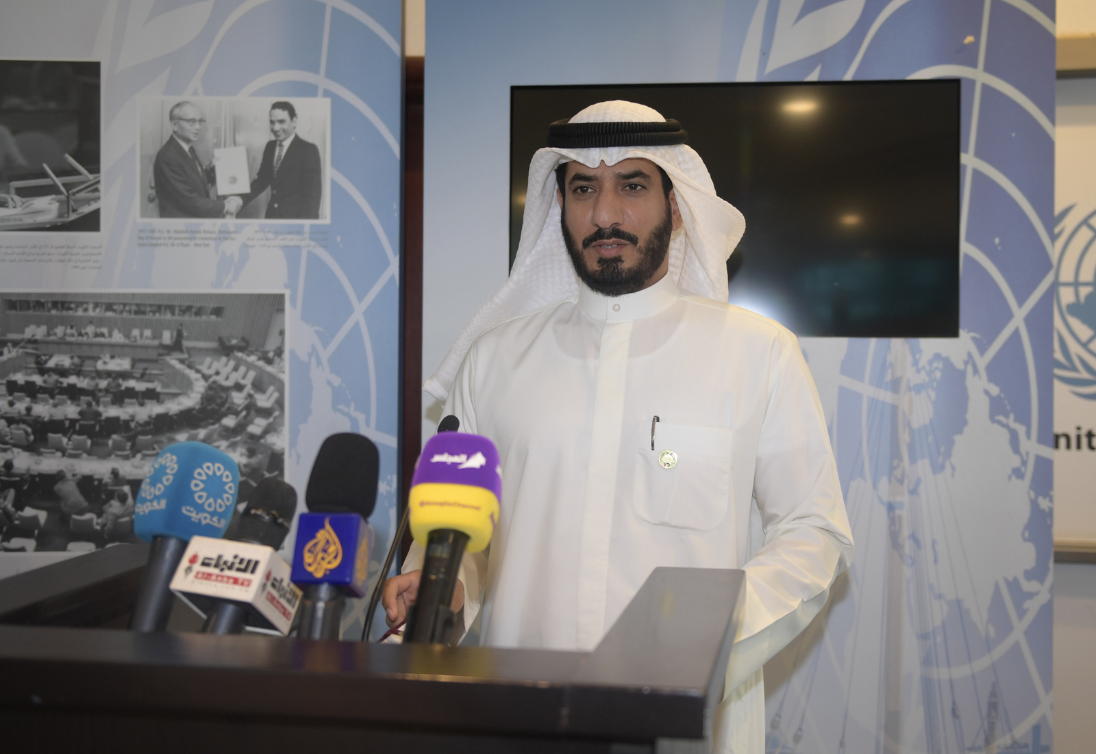Mr. Majed AlAzmi at a lectern with many microphones. There is a blank TV and UNHCR historic documents behind him.