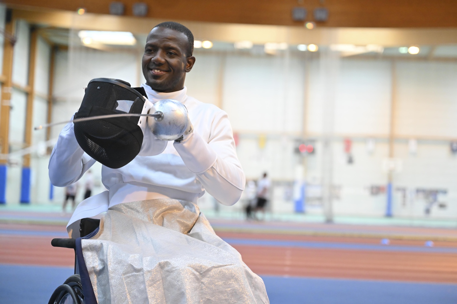 A man sits in a wheelchair holding a fencing epee and mask.