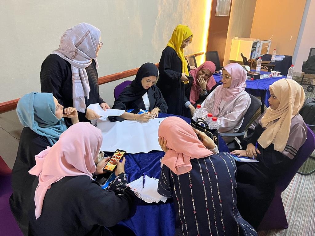 A group of women gather around a table at a training session, writing on large paper and looking at their phones