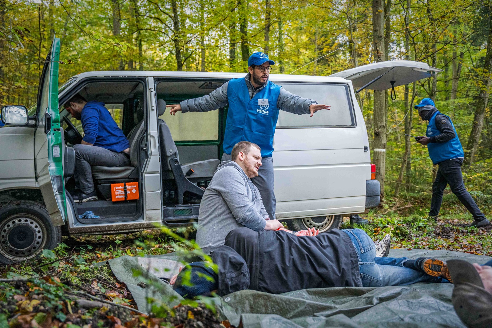 Five people participate in a training session in the forest. One sits on a tarp beside one laying down. One sits in a van with the doors and boot open, another stands in front of the van, pointing in two directions, and the fifth walks towards the van.