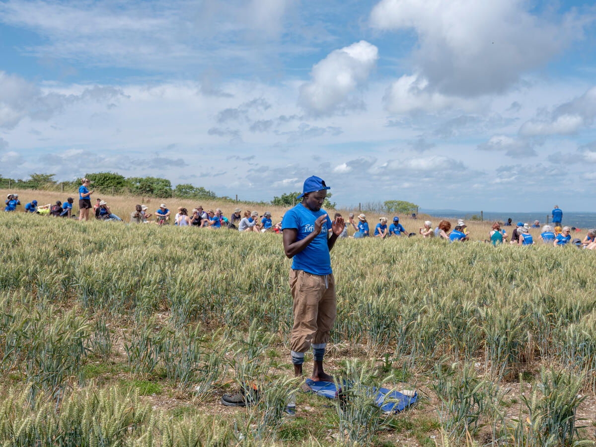 Man stands in field to perform prayers. People picnic behind him in the distance.