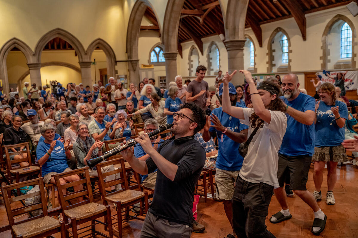 Clarinet player leads a column of dancing people around a church nave, as others, seated, watch.