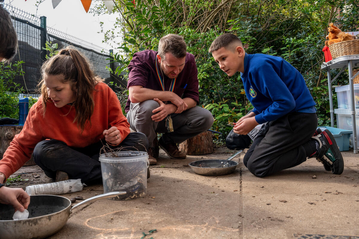 In a school playground a boy in school uniform leans over a frying pan whilst a male teacher supervises. A female teacher is at the front of the shot helping another pupil.