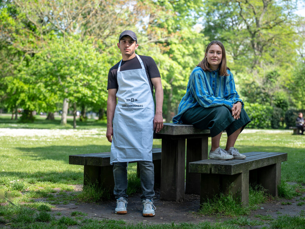 In a park, a woman sits on a picnic bench whilst a man stands.