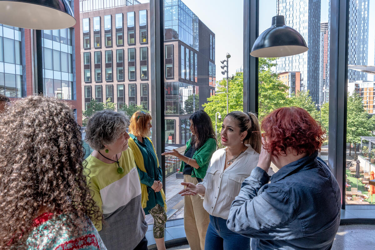 Seven women chat in a glass-walled room with buildings and trees seen in background. 