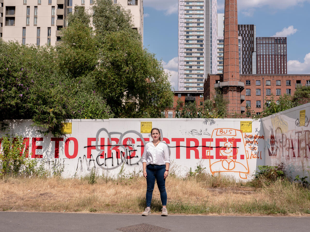 Woman poses stood in front of graffitied billboards reading “Welcome to First Street”, with skyscrapers in background.