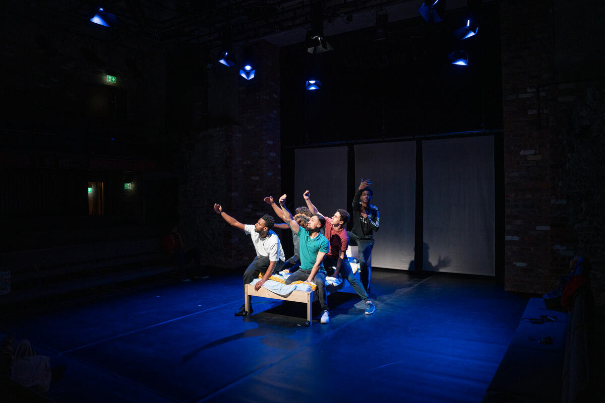 Six men perform in a play, kneeling on a bed.