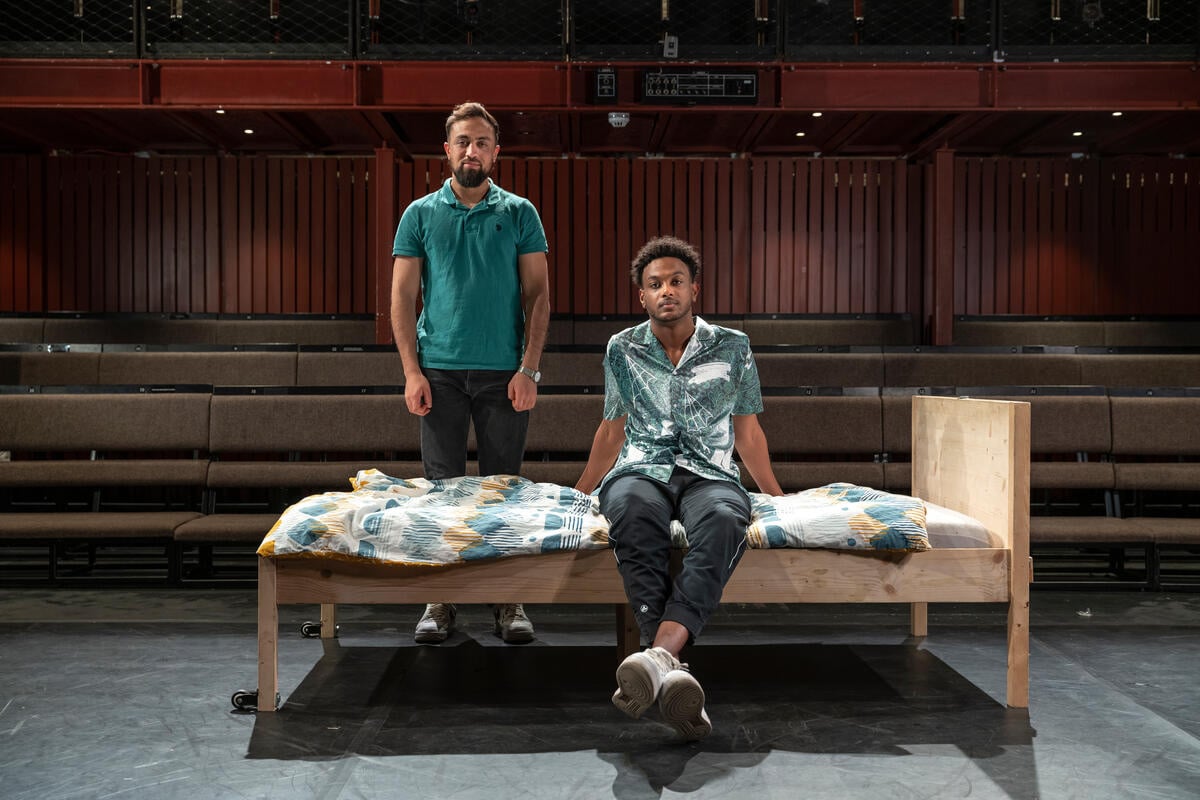 Two men pose in a theatre, one sitting on a bed, one standing behind.