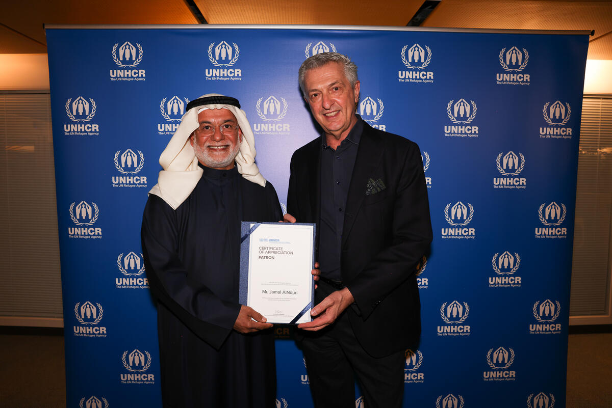 Two smiling men holding a certificate of appreciation, with a UNHCR banner behind them