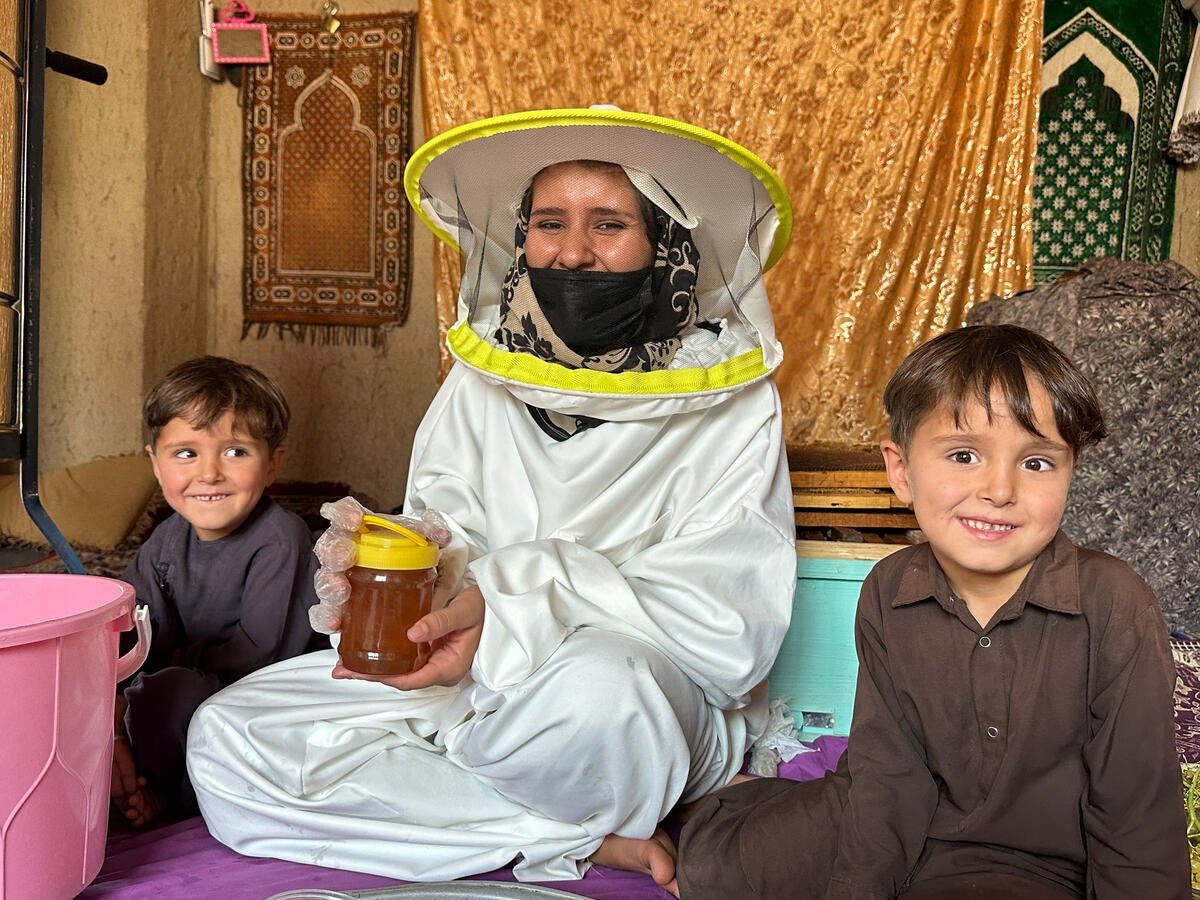 A woman wearing a white protective suit showcases a jar of honey while her children sit next to her inside their house