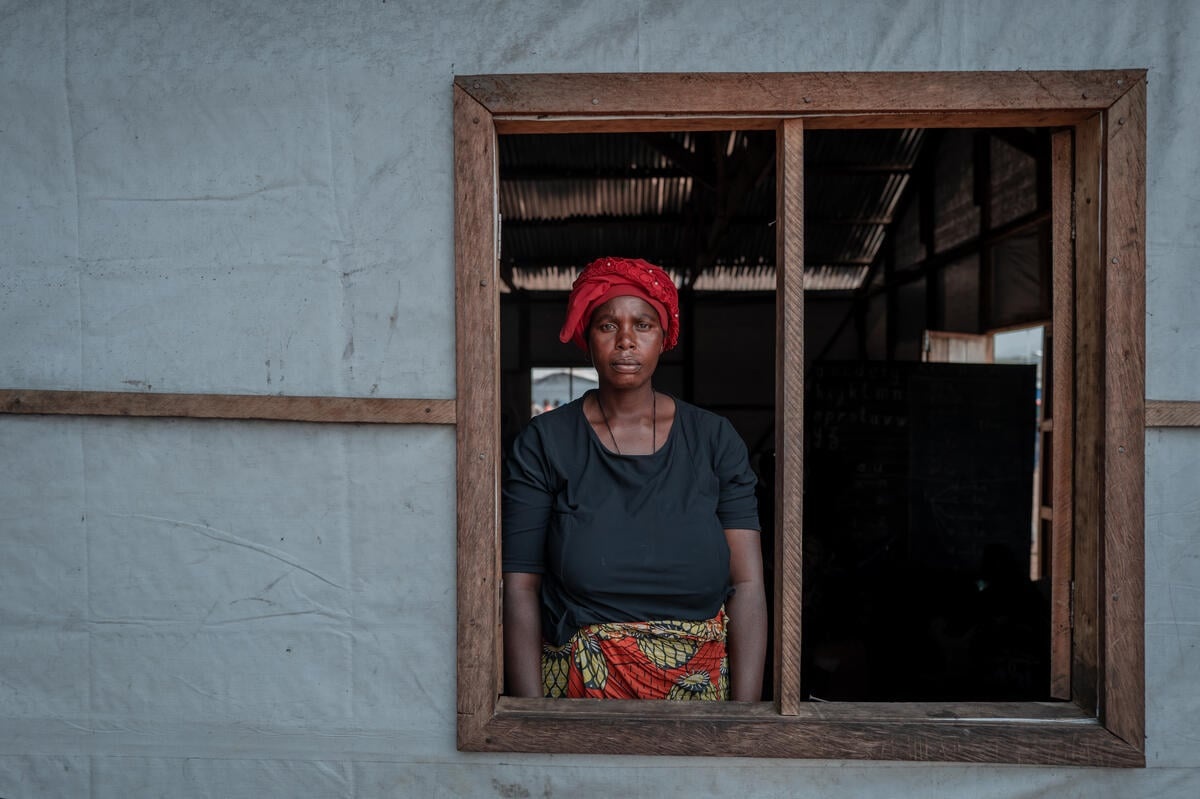 A woman wearing a red headscarf and black t-shirt looks out of an open wood-framed window set in the white plastic-sheet wall of a shelter. 
