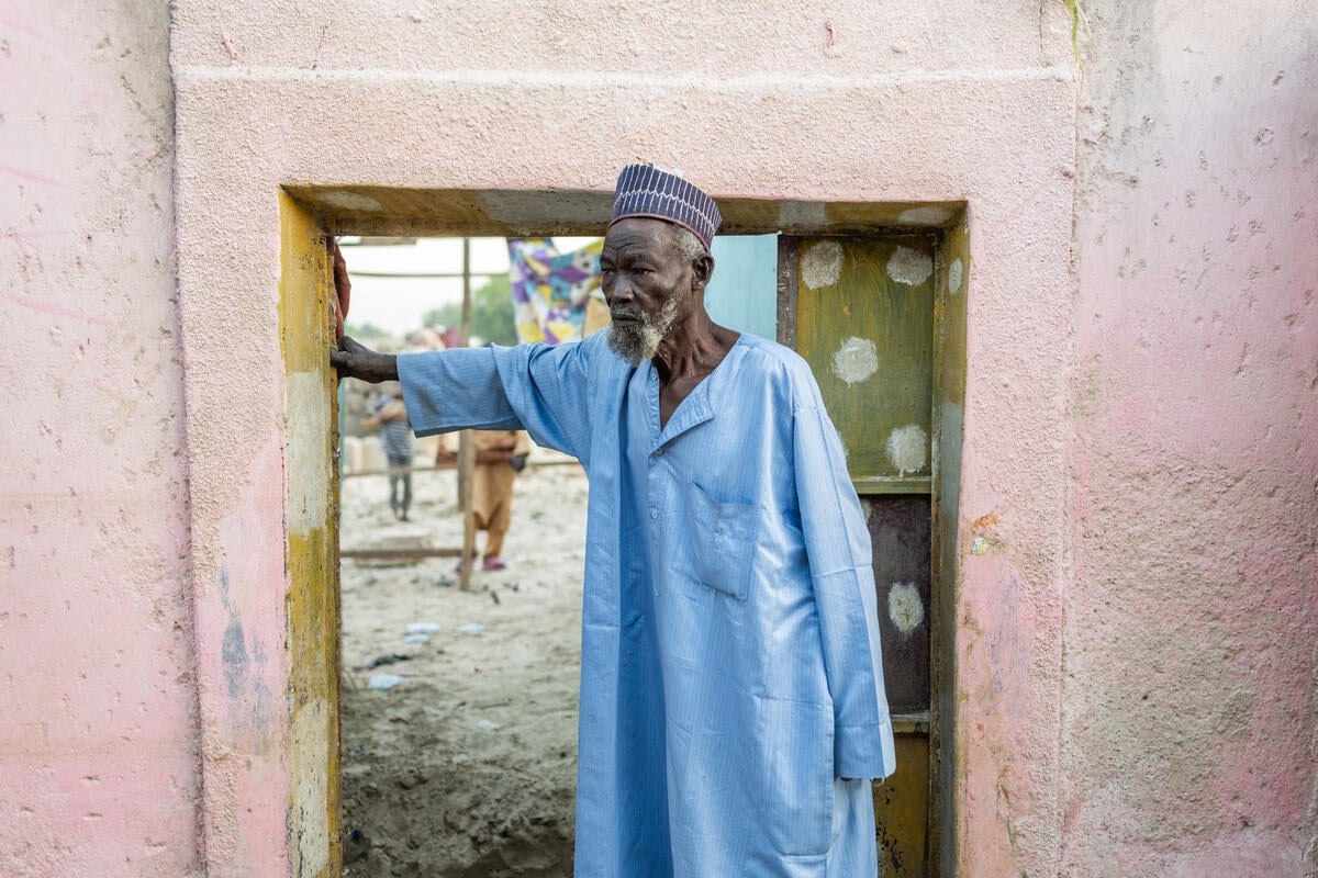 An old man stands in the doorway of a damaged house.