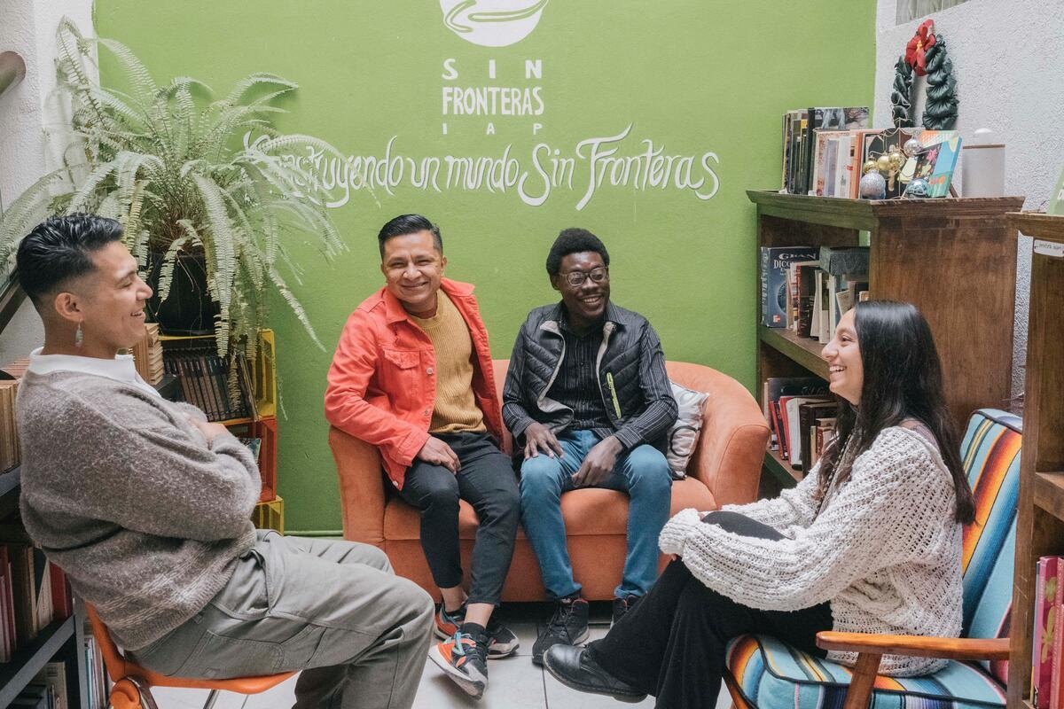 Two men sit on a small orange couch talking to a man and a woman sitting on chairs in a room lined with book cases.