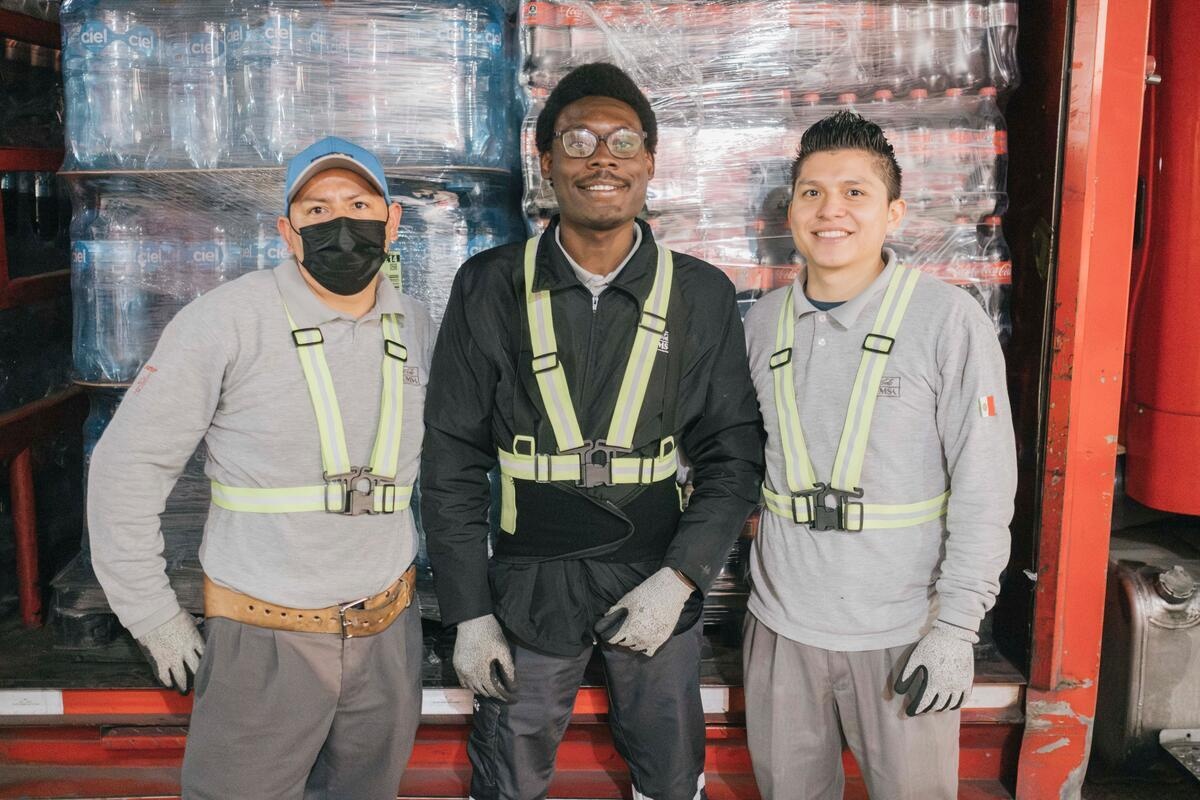 Three men pose at the back of a delivery truck containing soft drinks.