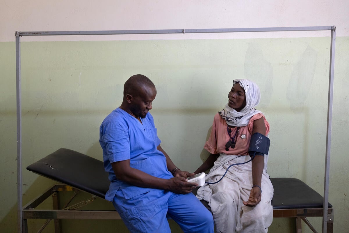 A male nurse wearing blue scrubs sits on a hospital bed next to a woman while he measures her blood pressure