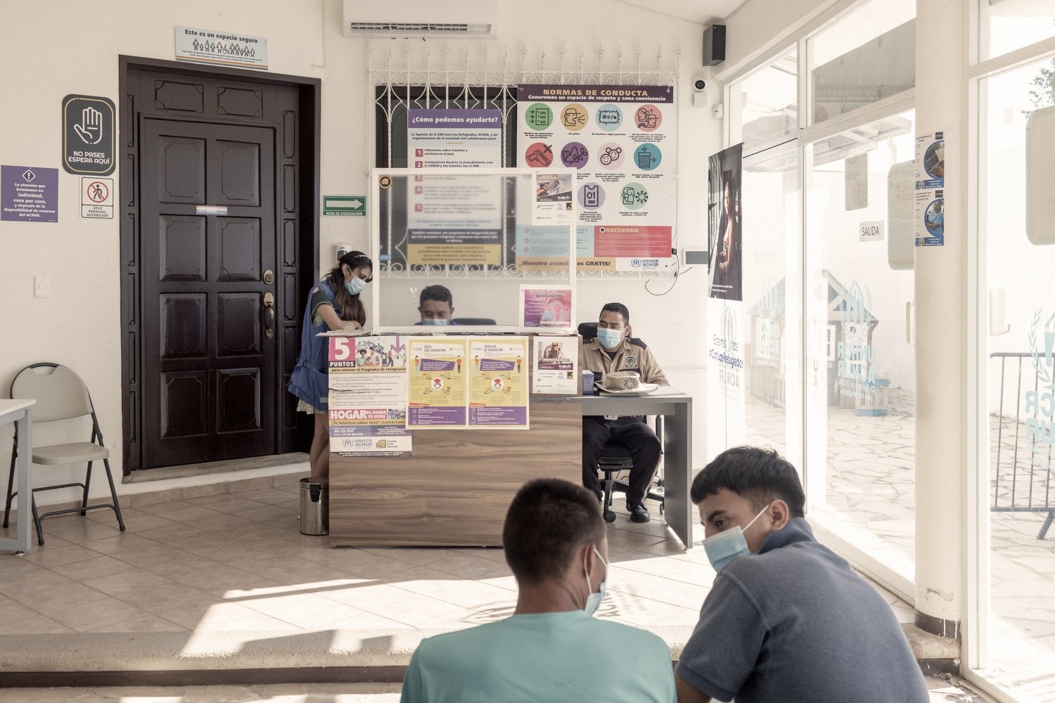 Two men wait to be assisted while officials sit behind a desk.