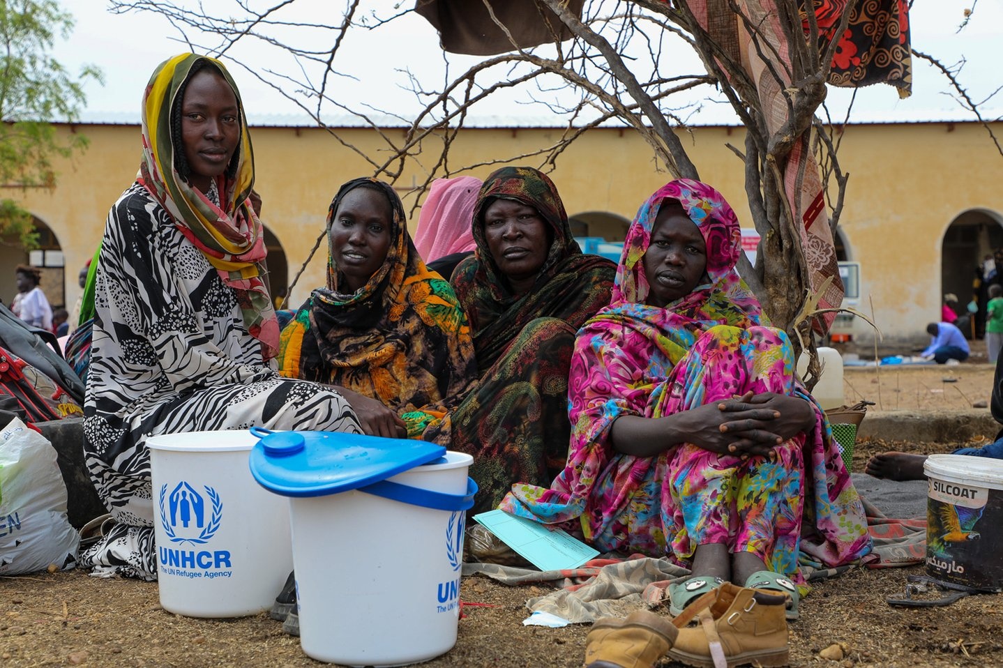 Four women sit on the ground outside a transit centre in Renk, South Sudan, with two UNHCR-branded buckets.