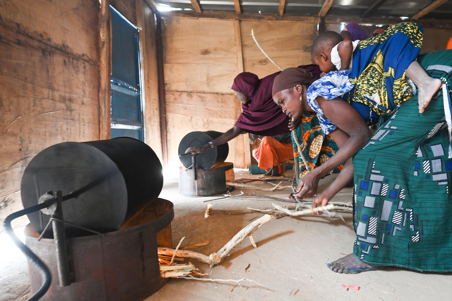 Two women stoke two wood-burning stoves in a shed-like structure.