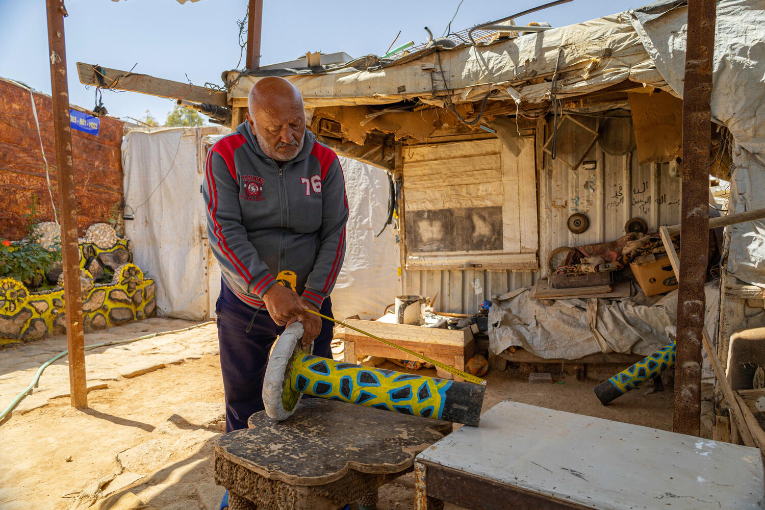 A man measures a colourful object outside his shelter.