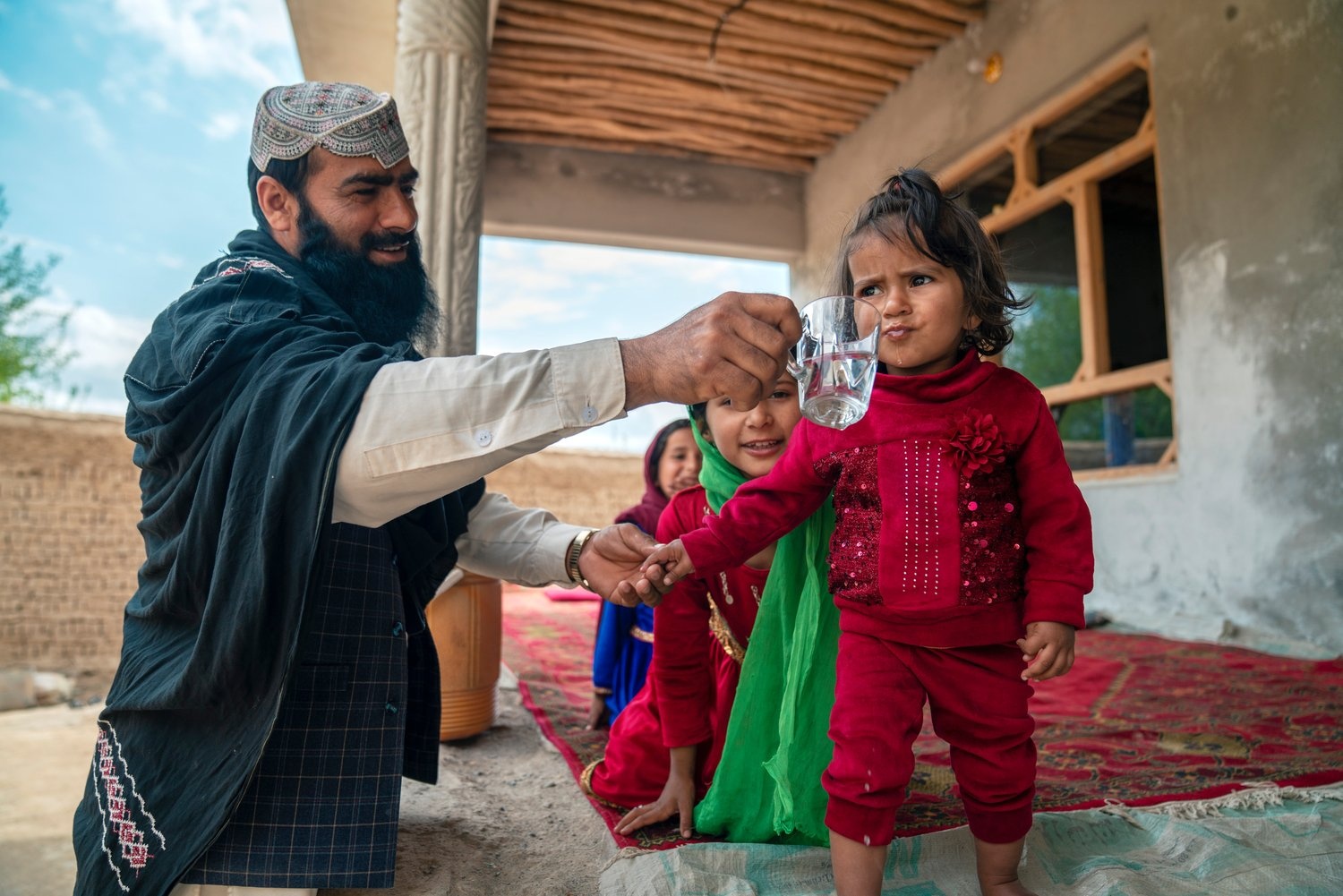 A man smiles as he holds a glass of water for a young girl to drink from.