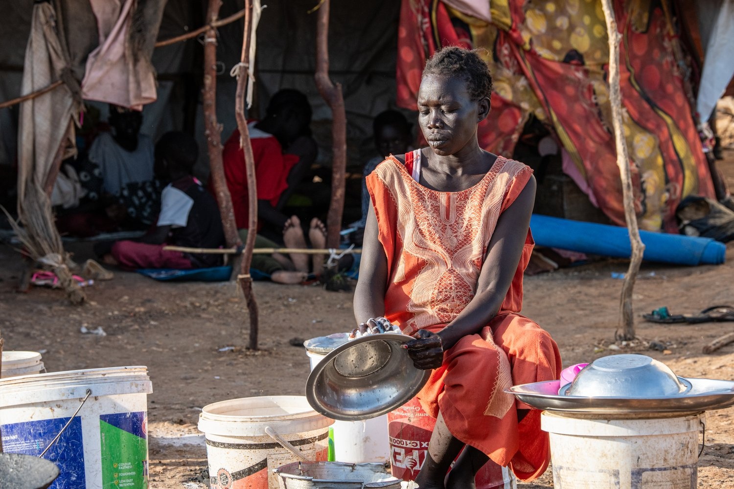 A woman sits on a bucket washing dishes outside a makeshift shelter.