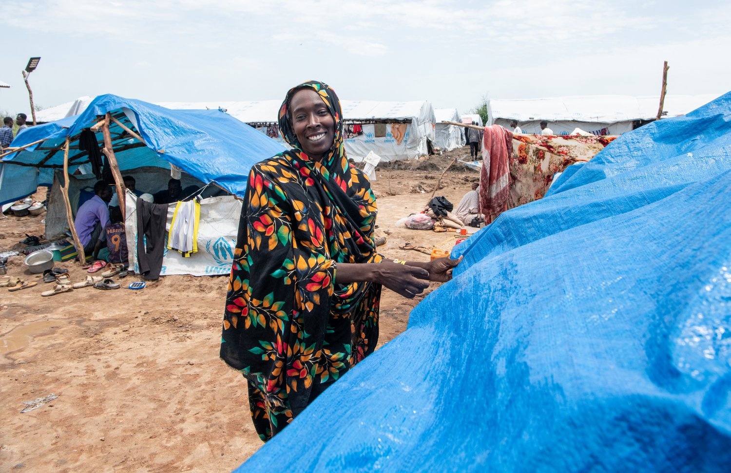 A woman smiles as she pulls a blue tarpaulin into place over a makeshift shelter.