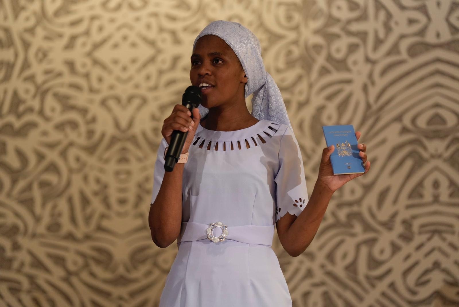 A woman on stage in front of a patterned backdrop speaks into a microphone while holding up a blue passport