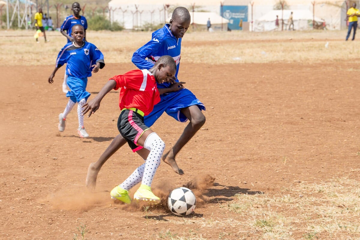 A smaller boy in a red jersey battles for the football against a taller player in blue, with several other blue players behind.