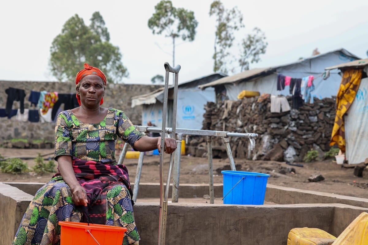 An elderly woman sits next to a communal tap holding a crutch in one hand and a bucket in the other.