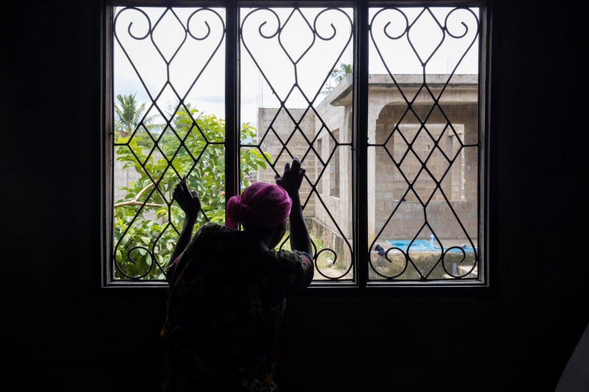 A woman with her back to the camera looks out of a window with her hands on the security bars.