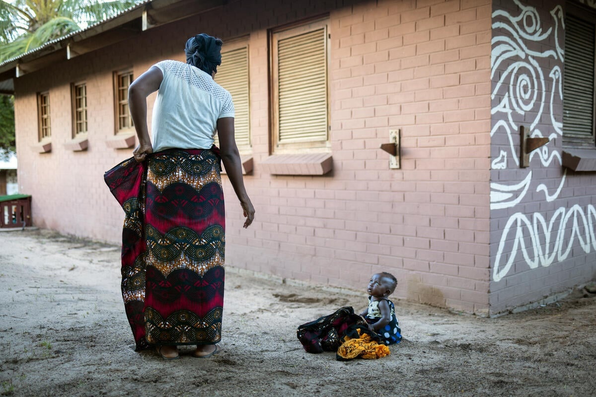 A woman adjusts her wrap-around skirt as a baby sitting on the ground looks on.