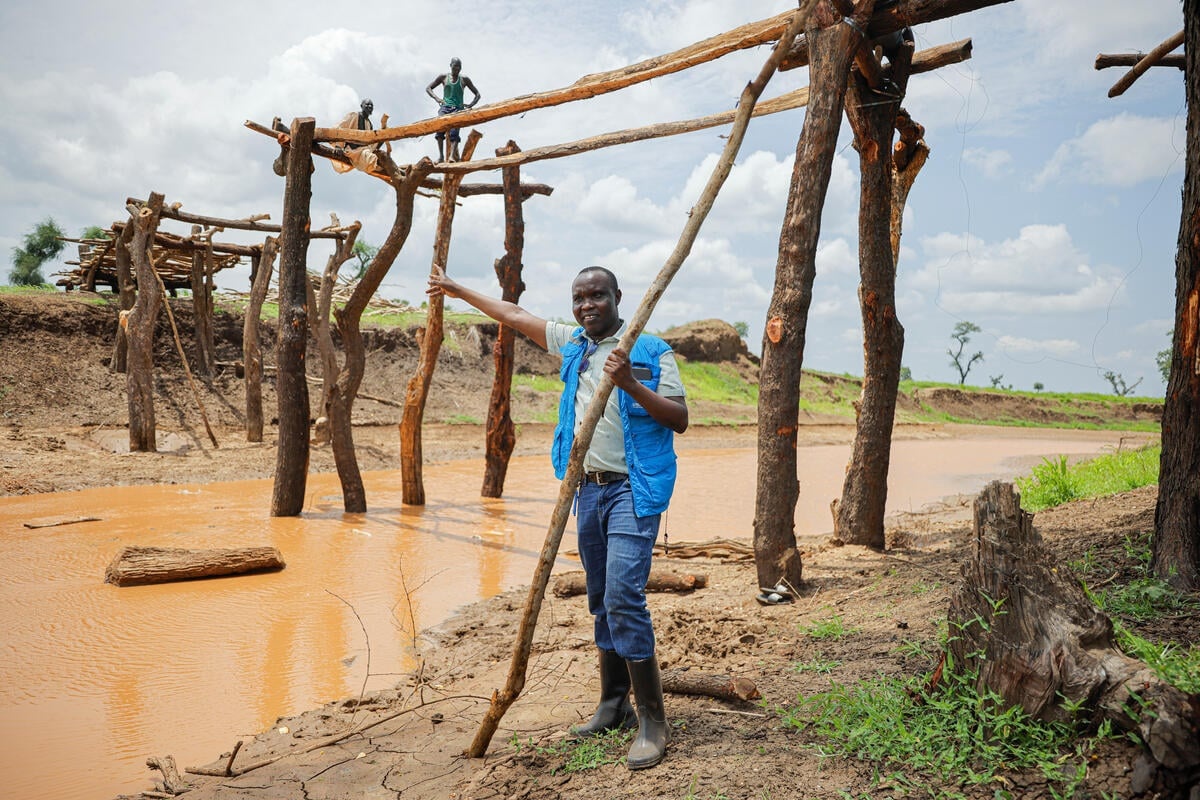 A man in a blue UNHCR vest stands under a footbridge being constructed over a river.
